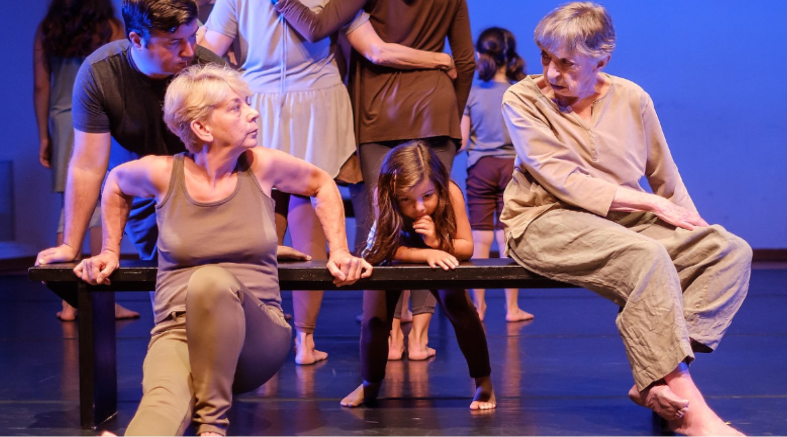 On a dance floor, under stage lighting, two older women are at opposite ends of a bench with a small child between them. The lady on the left is dipping her body in front of the bench with her hands and arms behind her, she looks directly into the eyes of the other lady who sits on the bench. The little girl stands behind the bench leaning on it with her elbows.